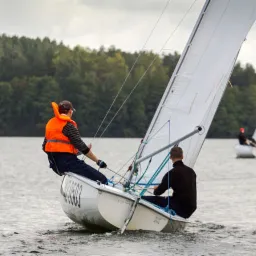 Dwaj mężczyźni na małej żaglówce podczas rejsu po jeziorze, jeden w kamizelce ratunkowej, w tle zalesiony brzeg i druga żaglówka.