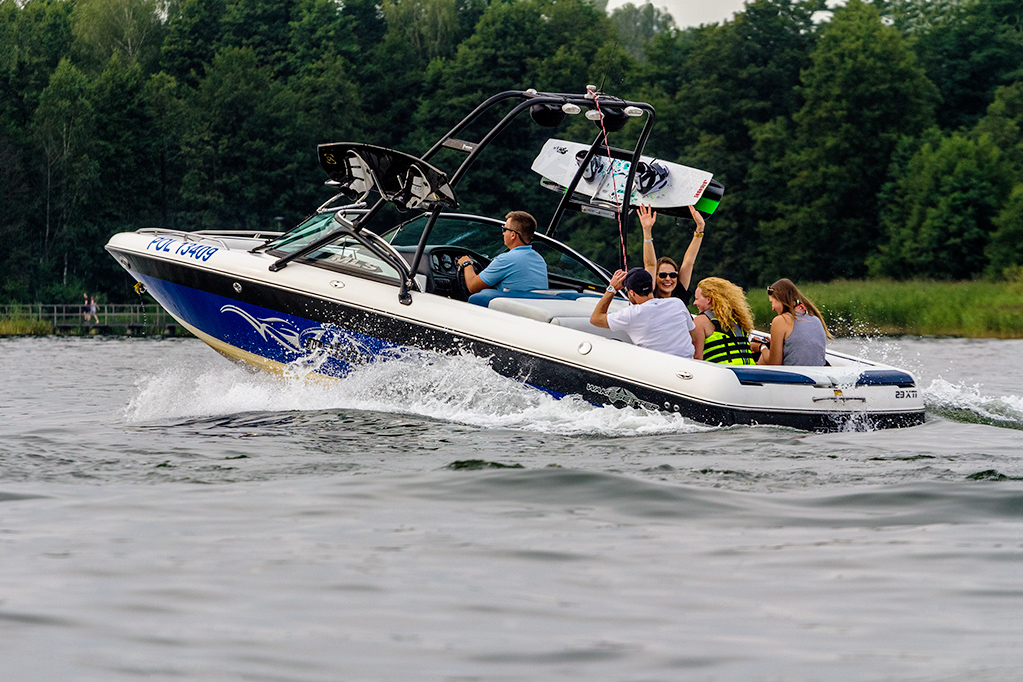 Motorówka z pasażerami płynie po jeziorze, wakeboard zamocowany na wieżyczce. Osoba z tyłu trzyma drugi wakeboard nad głową.