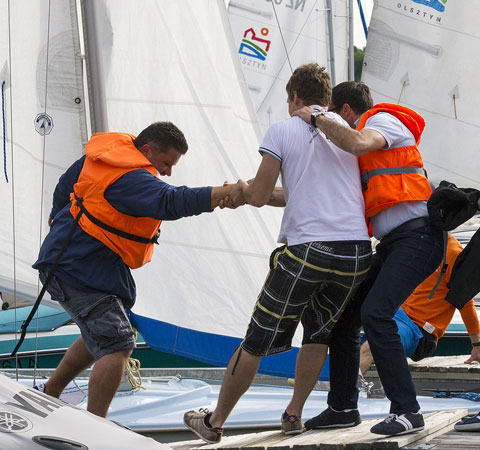 Grupa mężczyzn w kamizelkach ratunkowych pomaga wsiąść do żaglówki, widoczne białe żagle i nabrzeże.