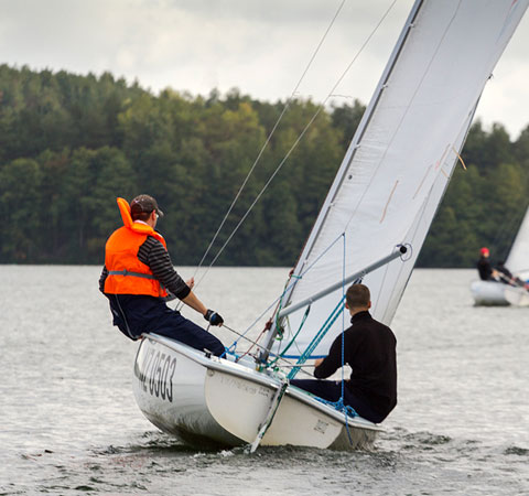 Dwaj mężczyźni na małej żaglówce podczas rejsu po jeziorze, jeden w kamizelce ratunkowej, w tle zalesiony brzeg i druga żaglówka.