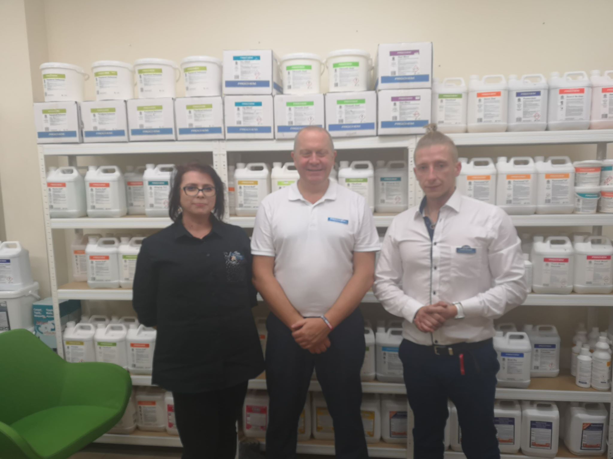 Trzy osoby stojące przed półkami z pojemnikami środków czystości marki Prochem, kobieta w czarnej koszuli, mężczyzna w białej koszulce polo i mężczyzna w białej koszuli z rozpiętym guzikiem...