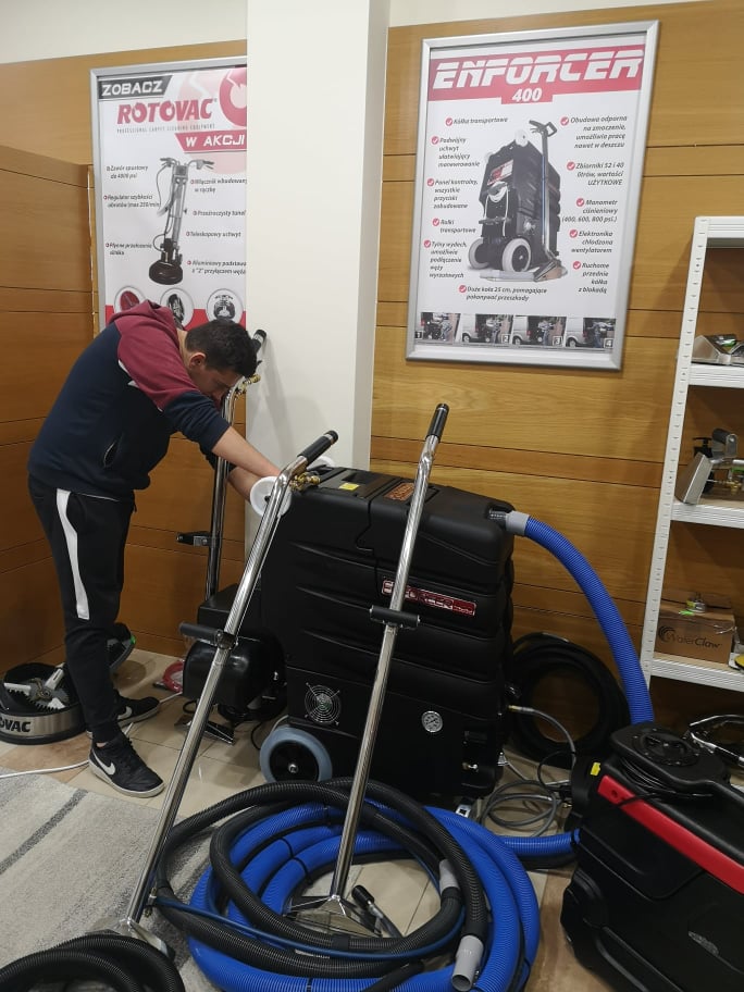 Mężczyzna w trakcie podłączania węża do profesjonalnego odkurzacza piorącego Enforcer 400, obok zwinięte węże i plakaty reklamowe urządzeń do czyszczenia dywanów.