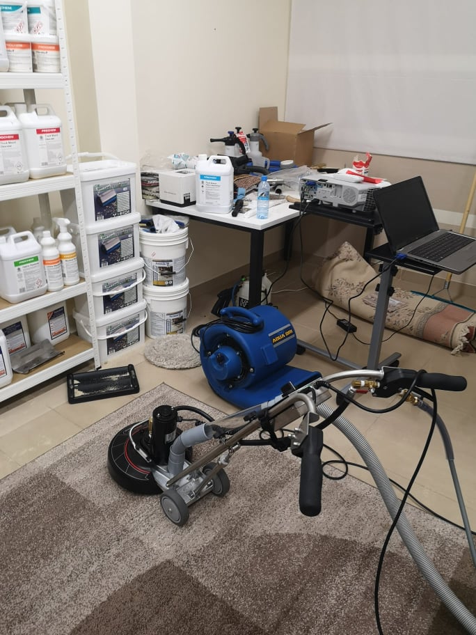 Maszyna do czyszczenia dywanów z szarym uchwytem i niebieską dmuchawą w tle, obok półki z pojemnikami środków czystości i stołu z akcesoriami.
