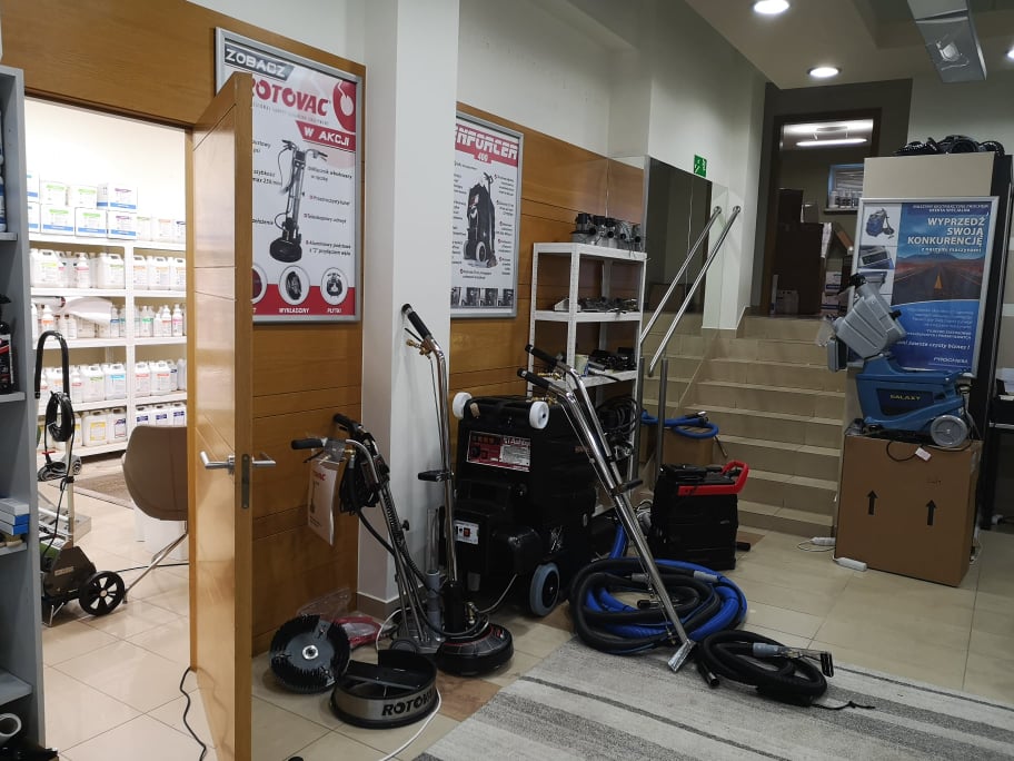 Wnętrze sklepu z maszynami do czyszczenia i środkami czystości, widoczne urządzenia do prania dywanów, polerki i półki z pojemnikami. Na ścianach plakaty reklamowe.