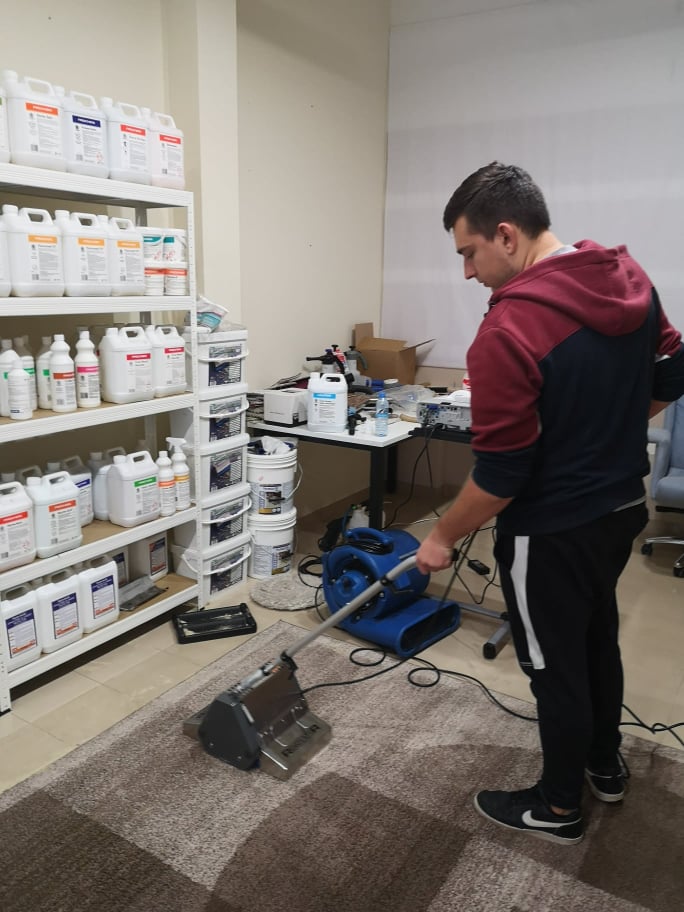 Mężczyzna czyści dywan za pomocą profesjonalnego odkurzacza piorącego; na półkach w tle widoczne środki czystości, a obok stoi dmuchawa do osuszania.