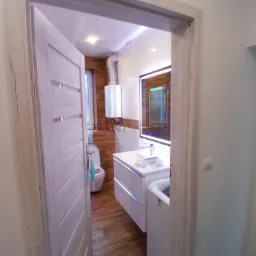 Wykończona łazienka z drewnopodobnymi płytkami, podświetlanym lustrem, białą szafką umywalkową z szufladami z niebieskim podświetleniem LED, toaletą i pralką, widziana zza częściowo otwartych...