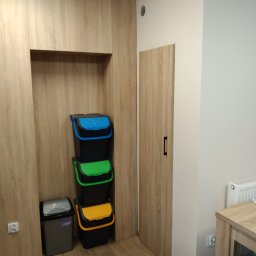 Dariusz Krzyżek - Szafa na wymiar z wnęką skrywającą kosze do segregacji odpadów: niebieski, zielony, żółty i szary. Drzwi szafy w kolorze drewna, pasujące do podłogi.