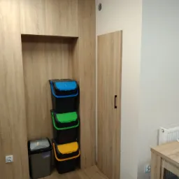 Szafa na wymiar z wnęką skrywającą kosze do segregacji odpadów: niebieski, zielony, żółty i szary. Drzwi szafy w kolorze drewna, pasujące do podłogi.