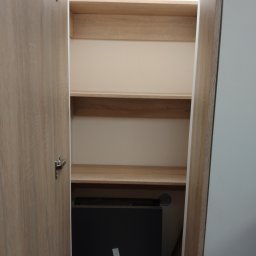 Dariusz Krzyżek - Otwarta szafa z jasnego drewna z trzema półkami i widocznym fragmentem czarnego urządzenia w dolnej części. Drzwi szafy z widocznym zawiasem.