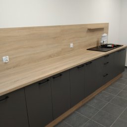 Dariusz Krzyżek - Nowoczesna, długa zabudowa kuchenna z blatem imitującym drewno i szarymi szafkami z czarnymi uchwytami. Widoczny zlew i gniazdka elektryczne na ścianie.