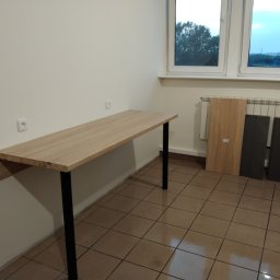 Dariusz Krzyżek - Blat biurka z dębowego drewna, wsparty na czarnych nogach, zamontowany do ściany. W tle okno i grzejnik z panelami. Widoczne gniazdka elektryczne na ścianie.