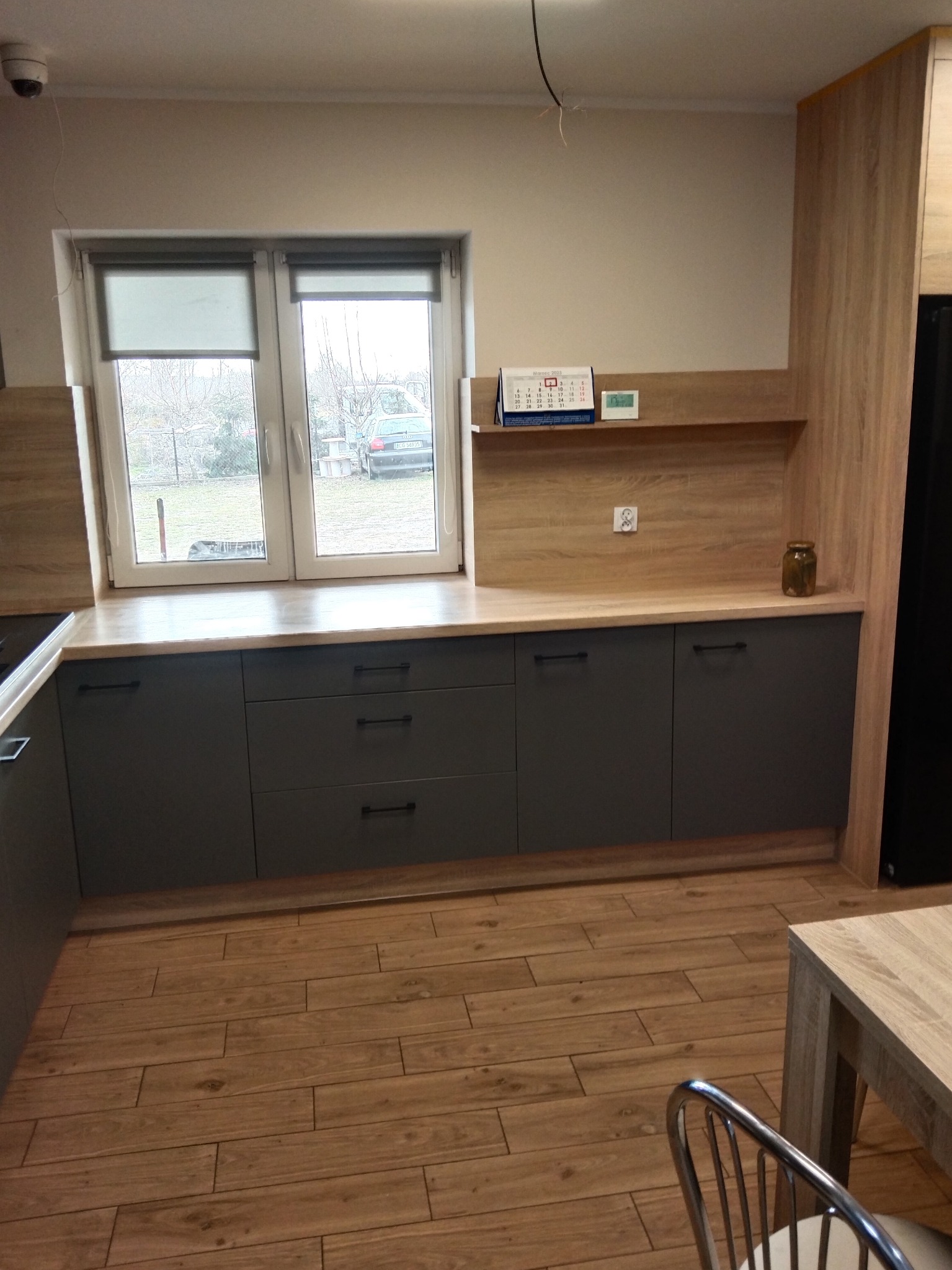 Szare szafki kuchenne z drewnianym blatem pod oknem z roletami. Na blacie kalendarz i słoik z ogórkami. Podłoga wyłożona drewnopodobnymi płytkami.