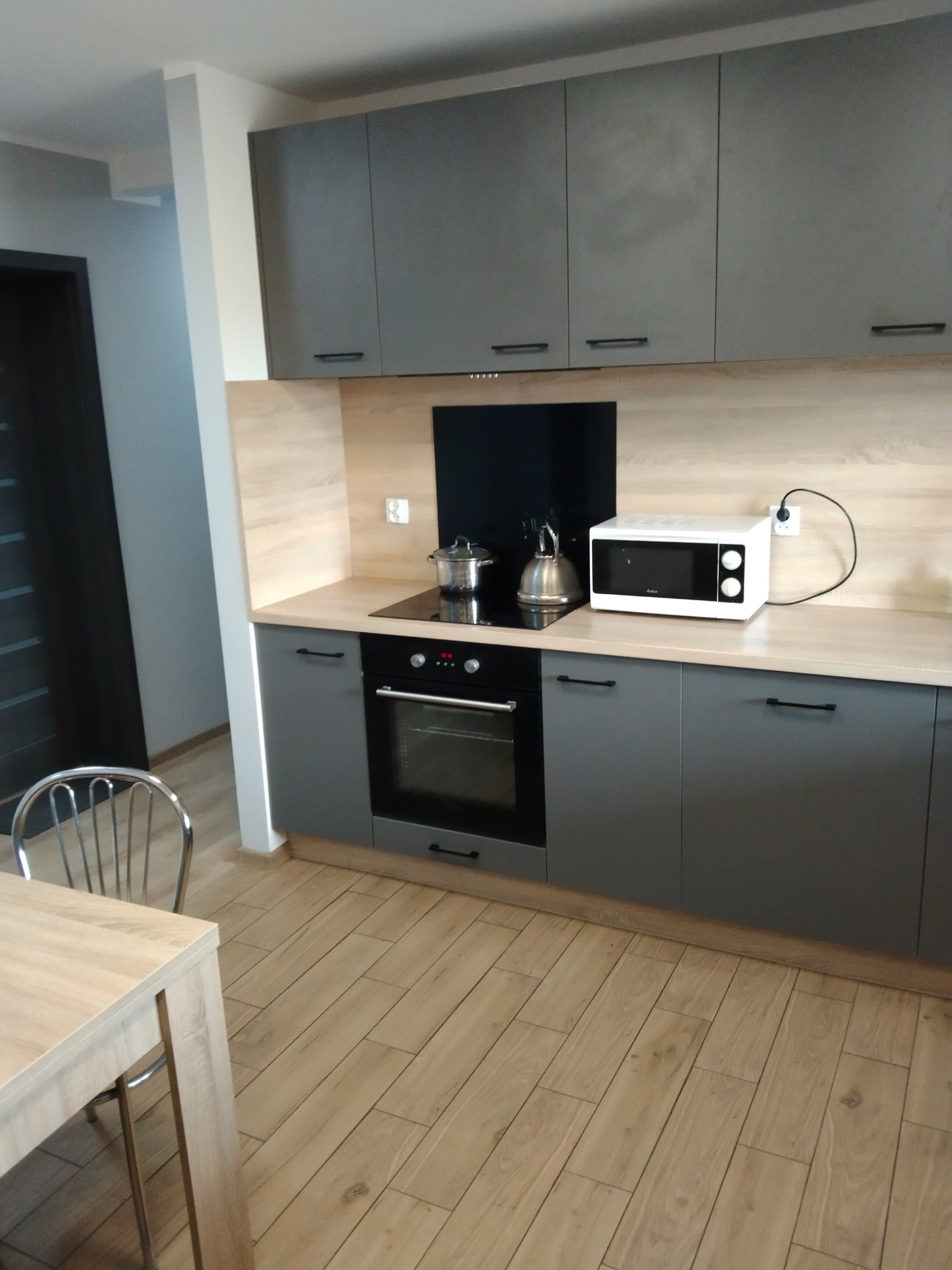 Szara zabudowa kuchenna z drewnianym blatem i czarnymi uchwytami. Na blacie czajnik, garnek i mikrofalówka. Podłoga wyłożona panelami imitującymi drewno.