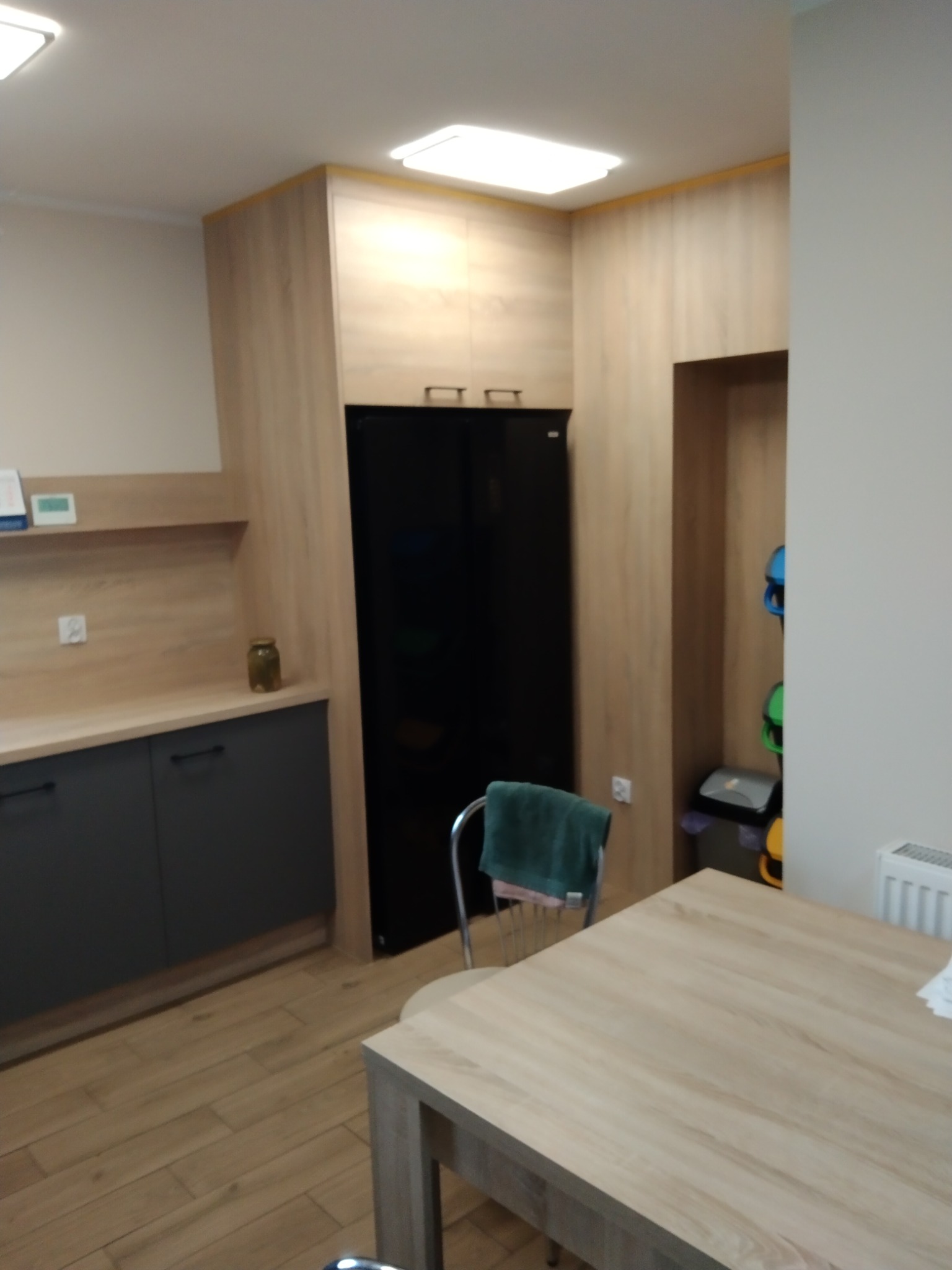 Nowoczesna kuchnia z drewnianymi szafkami i czarną lodówką. Stół z krzesłem w centrum, drewniana podłoga. Widoczne detale wykończenia.