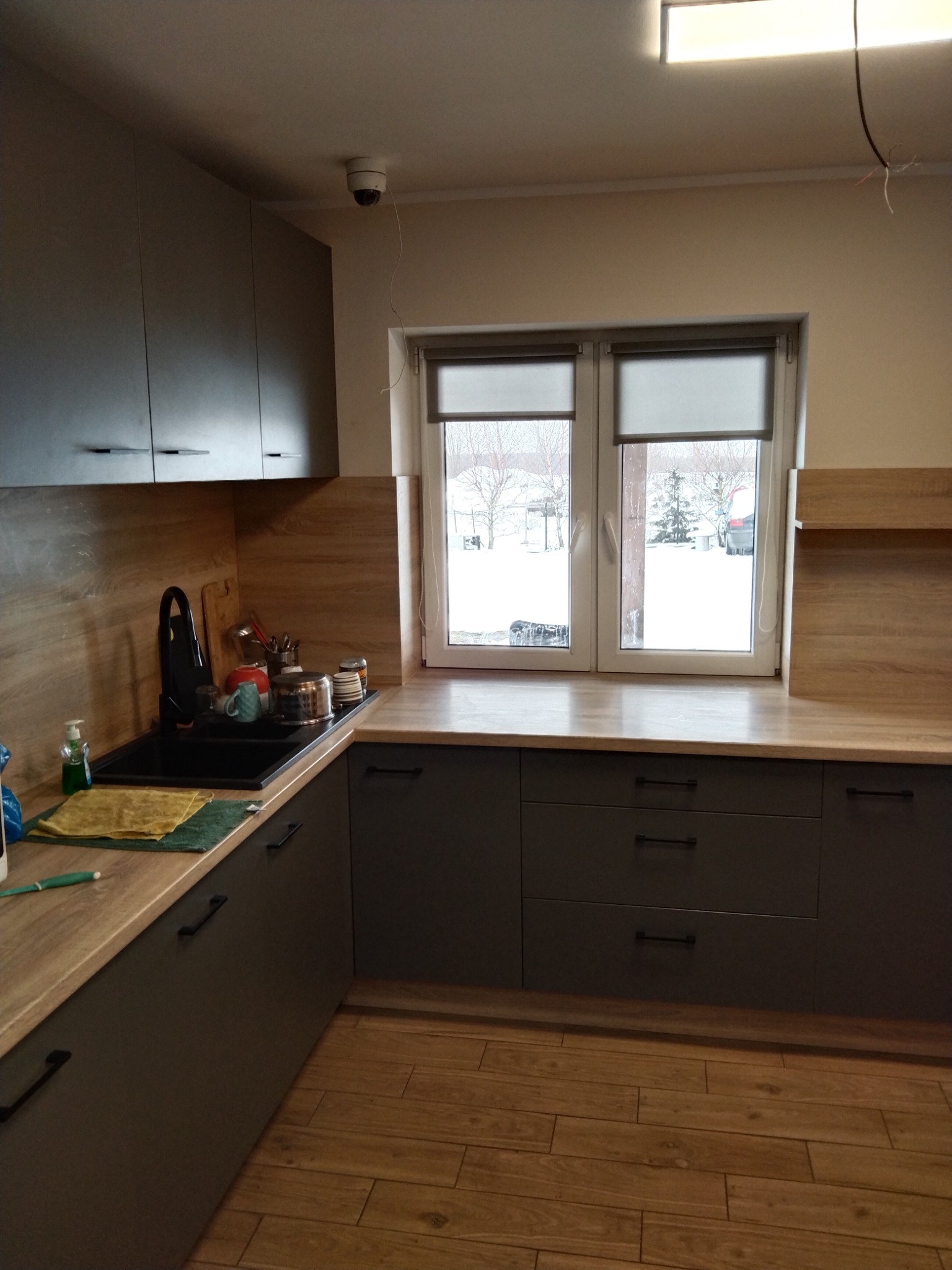Szara zabudowa kuchenna z drewnianym blatem i okapem, widok na okno z roletami i zimowy krajobraz. Umywalka z czarną baterią i akcesoriami. Nowoczesny design.