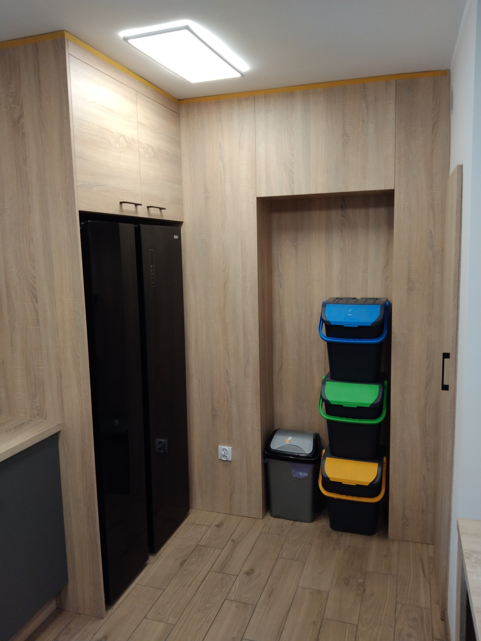 Funkcjonalna zabudowa kuchenna w Toruniu z drewnopodobnymi frontami, wkomponowana lodówka side-by-side i miejsce na sortowanie śmieci. Nowoczesny design.