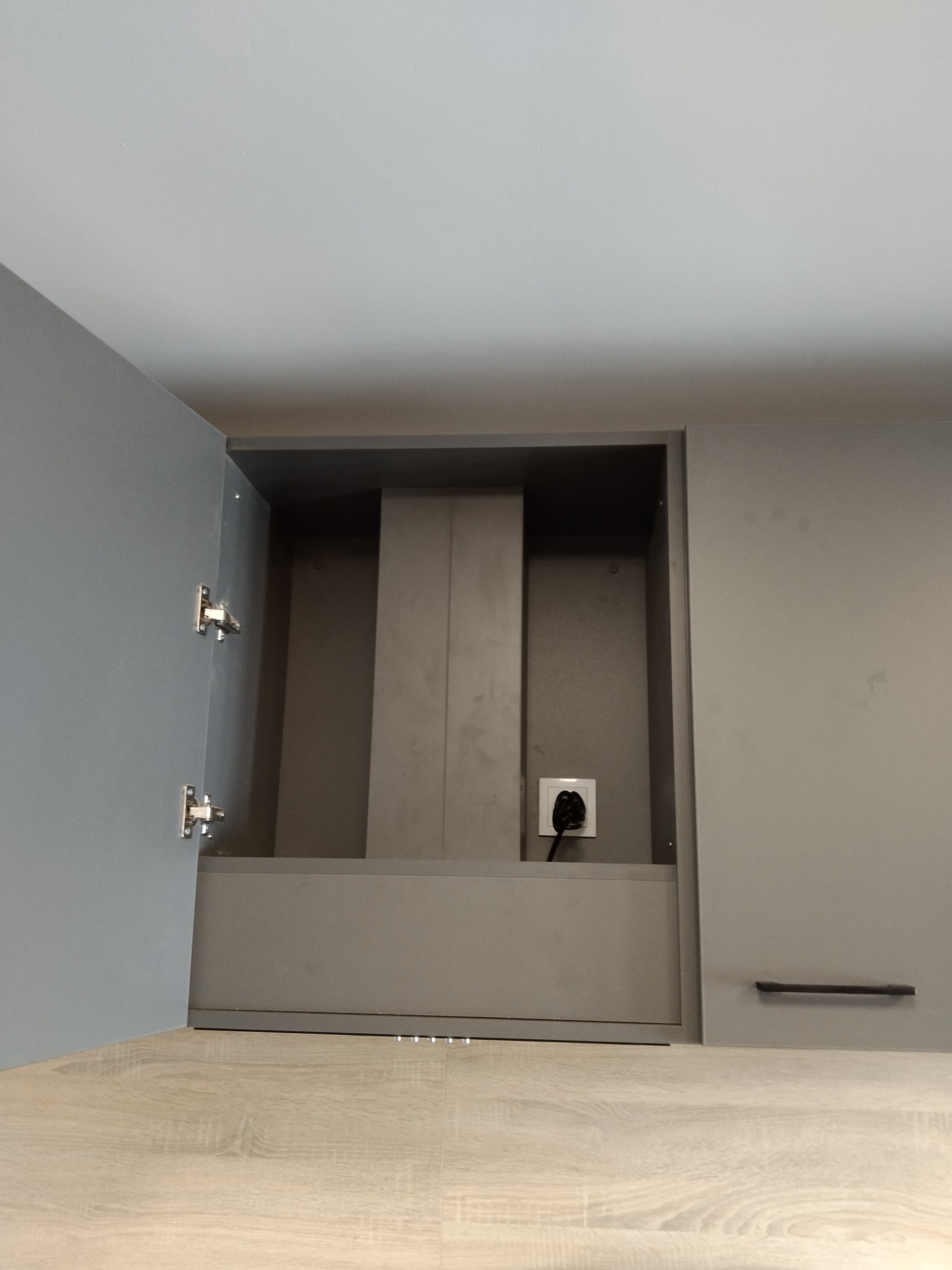 Szara zabudowa kuchenna z otwartą szafką, widoczny okap i gniazdko elektryczne. Ujęcie od dołu, podkreślające minimalistyczny design i funkcjonalność.