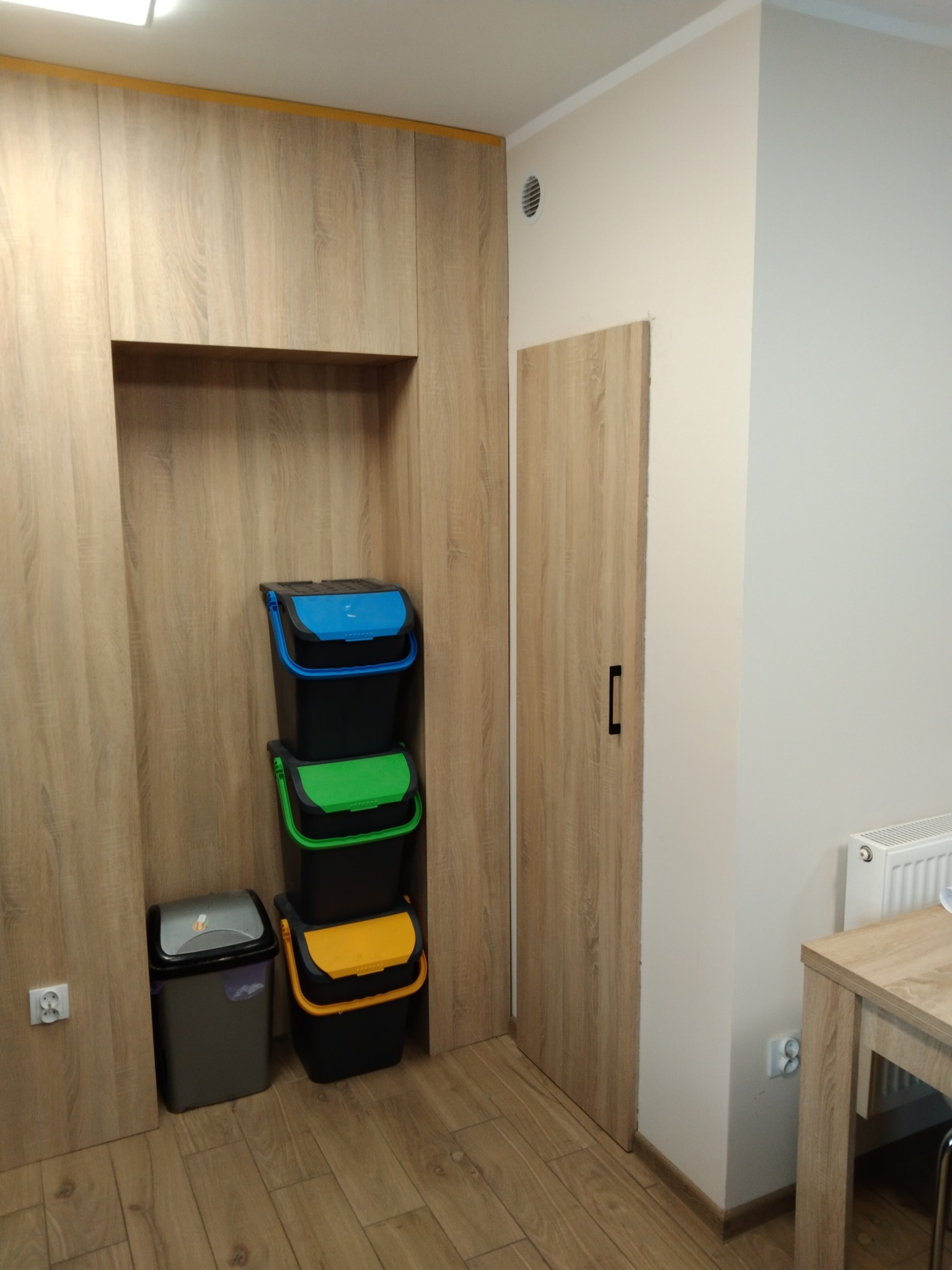 Szafa na wymiar z wnęką skrywającą kosze do segregacji odpadów: niebieski, zielony, żółty i szary. Drzwi szafy w kolorze drewna, pasujące do podłogi.