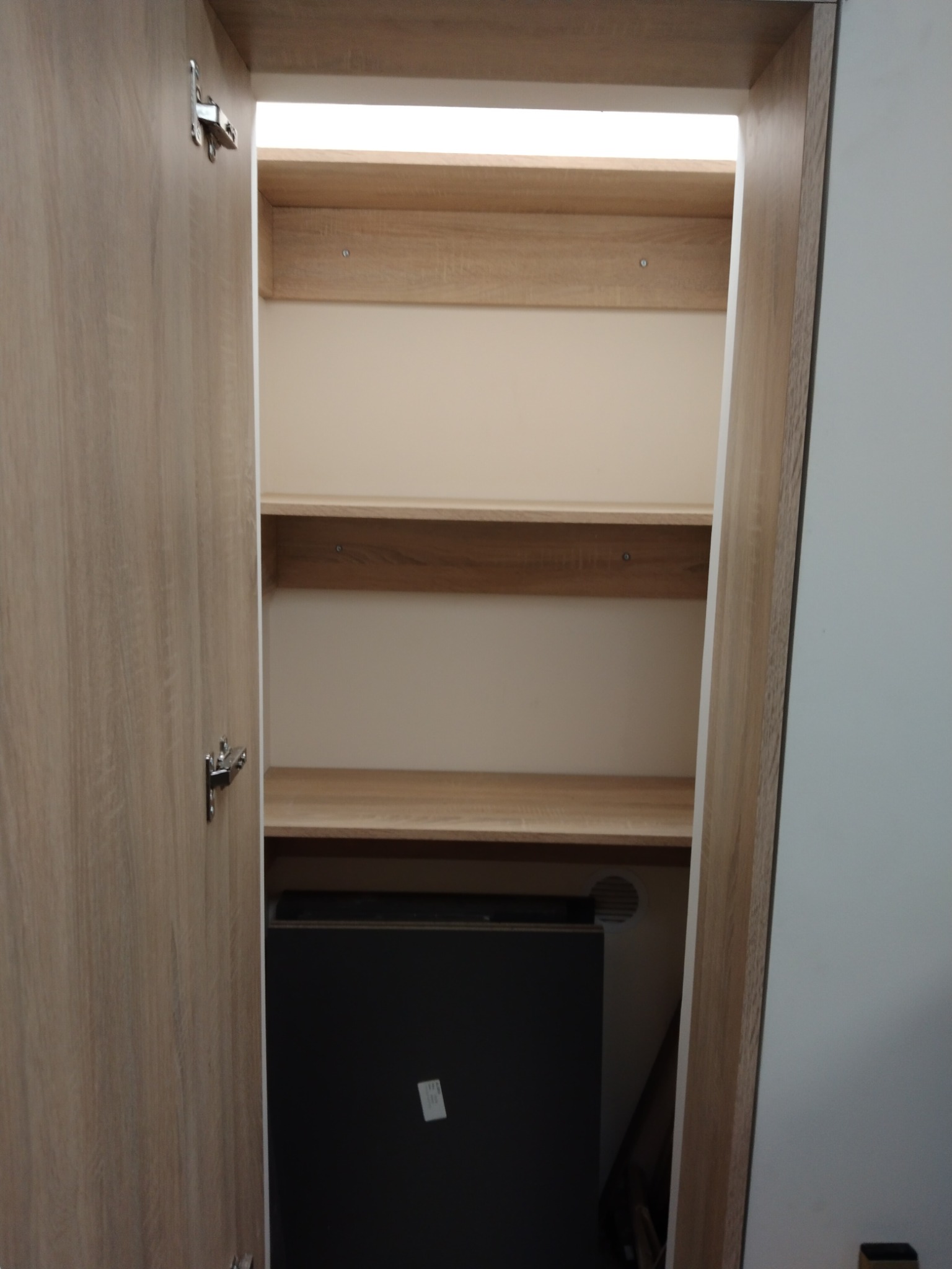 Otwarta szafa z jasnego drewna z trzema półkami i widocznym fragmentem czarnego urządzenia w dolnej części. Drzwi szafy z widocznym zawiasem.