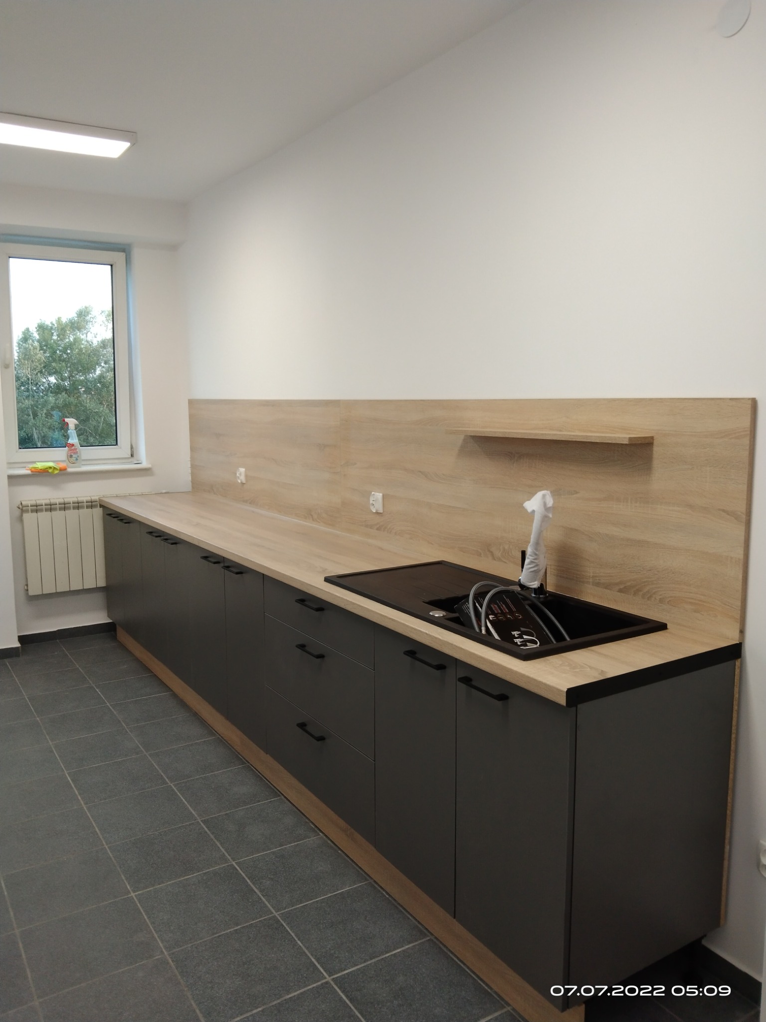 Nowoczesna kuchnia z ciemnymi szafkami i drewnianym blatem. Widoczny zlew z baterią i osprzętem. Minimalistyczny design, jasne ściany i szare płytki podłogowe.