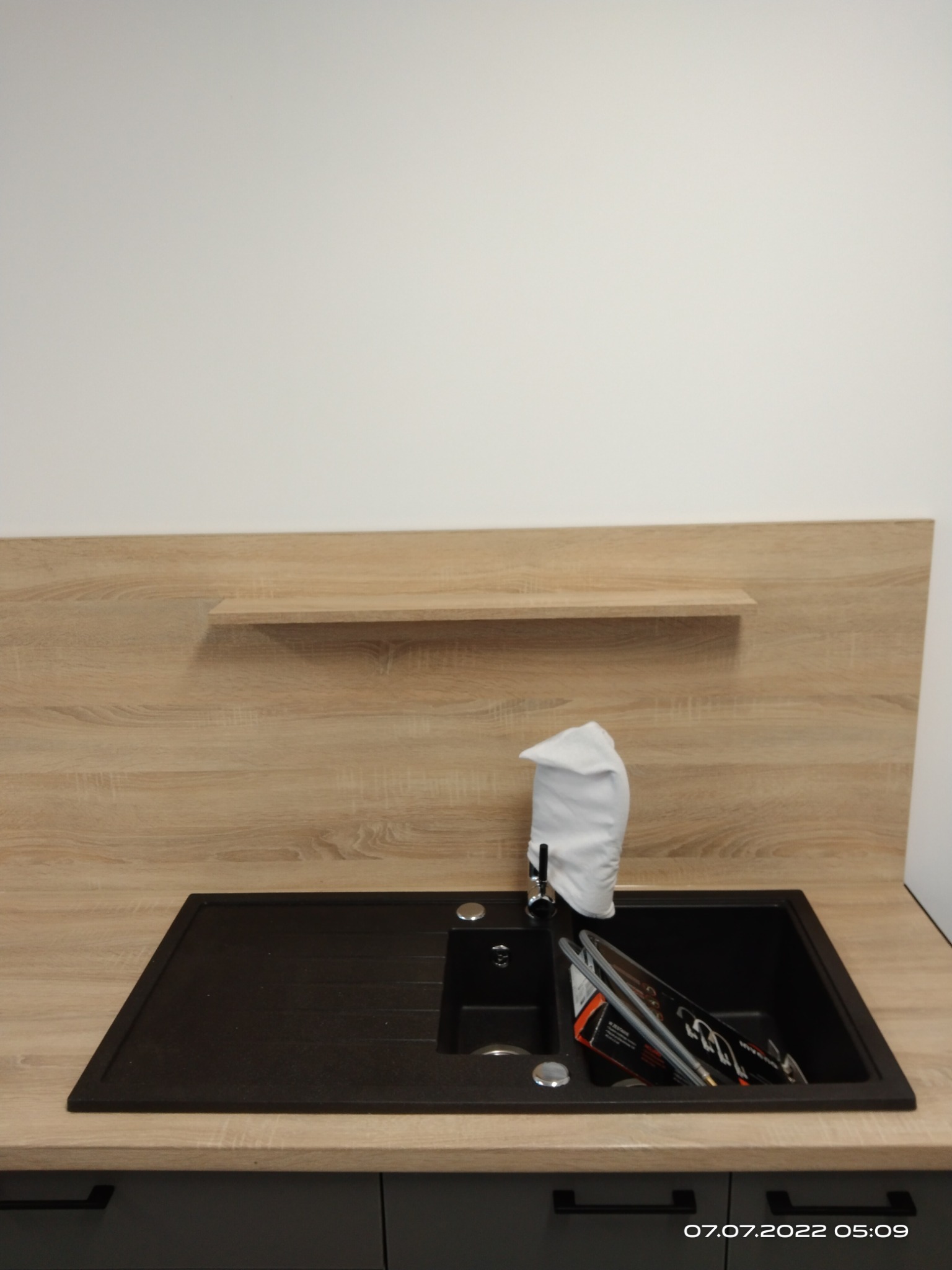 Nowoczesna, czarna, granitowa umywalka z baterią i drewnianym panelem ściennym z półką. Umywalka częściowo wypełniona akcesoriami. Widoczny fragment szafki.