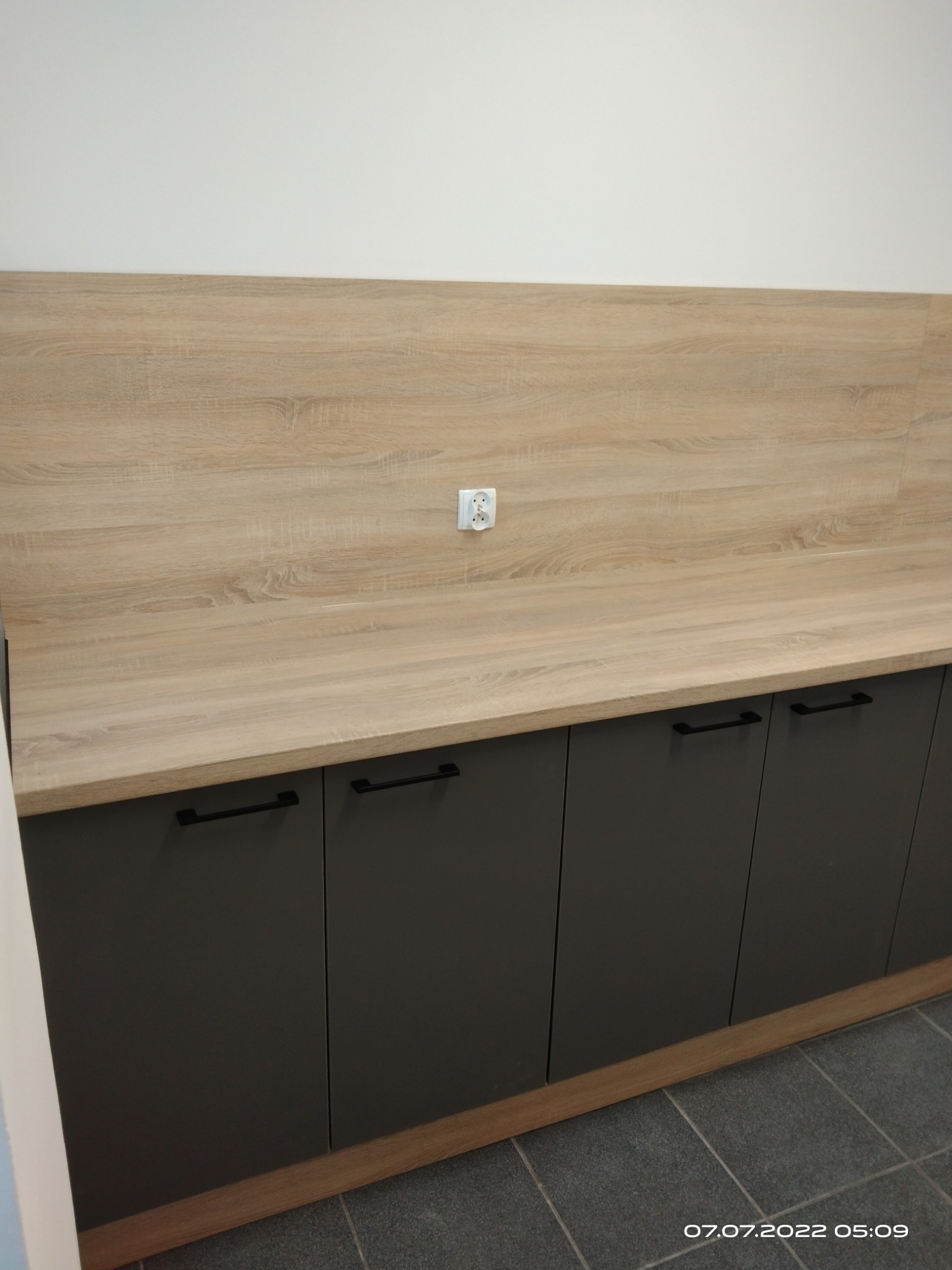 Nowoczesna zabudowa kuchenna z szarymi szafkami i blatem imitującym drewno. Widoczne czarne, minimalistyczne uchwyty i gniazdko elektryczne w blacie.