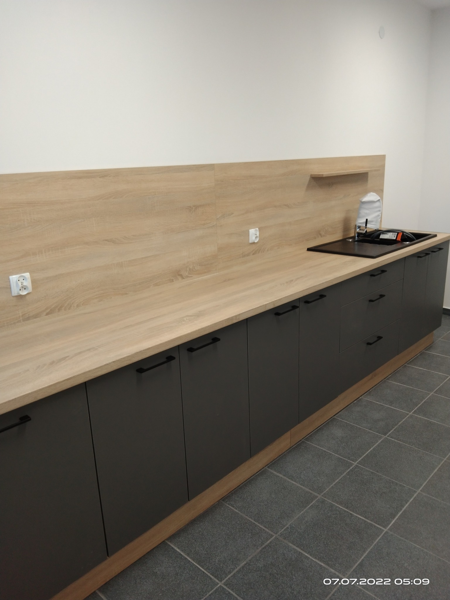 Nowoczesna, długa zabudowa kuchenna z blatem imitującym drewno i szarymi szafkami z czarnymi uchwytami. Widoczny zlew i gniazdka elektryczne na ścianie.