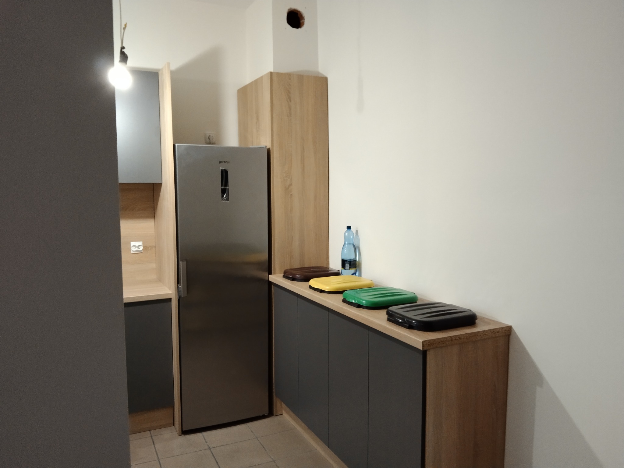 Nowoczesna kuchnia z szarymi szafkami i drewnianymi blatami. Na blacie pojemniki do segregacji śmieci. Widoczna lodówka ze stali nierdzewnej i wisząca żarówka.