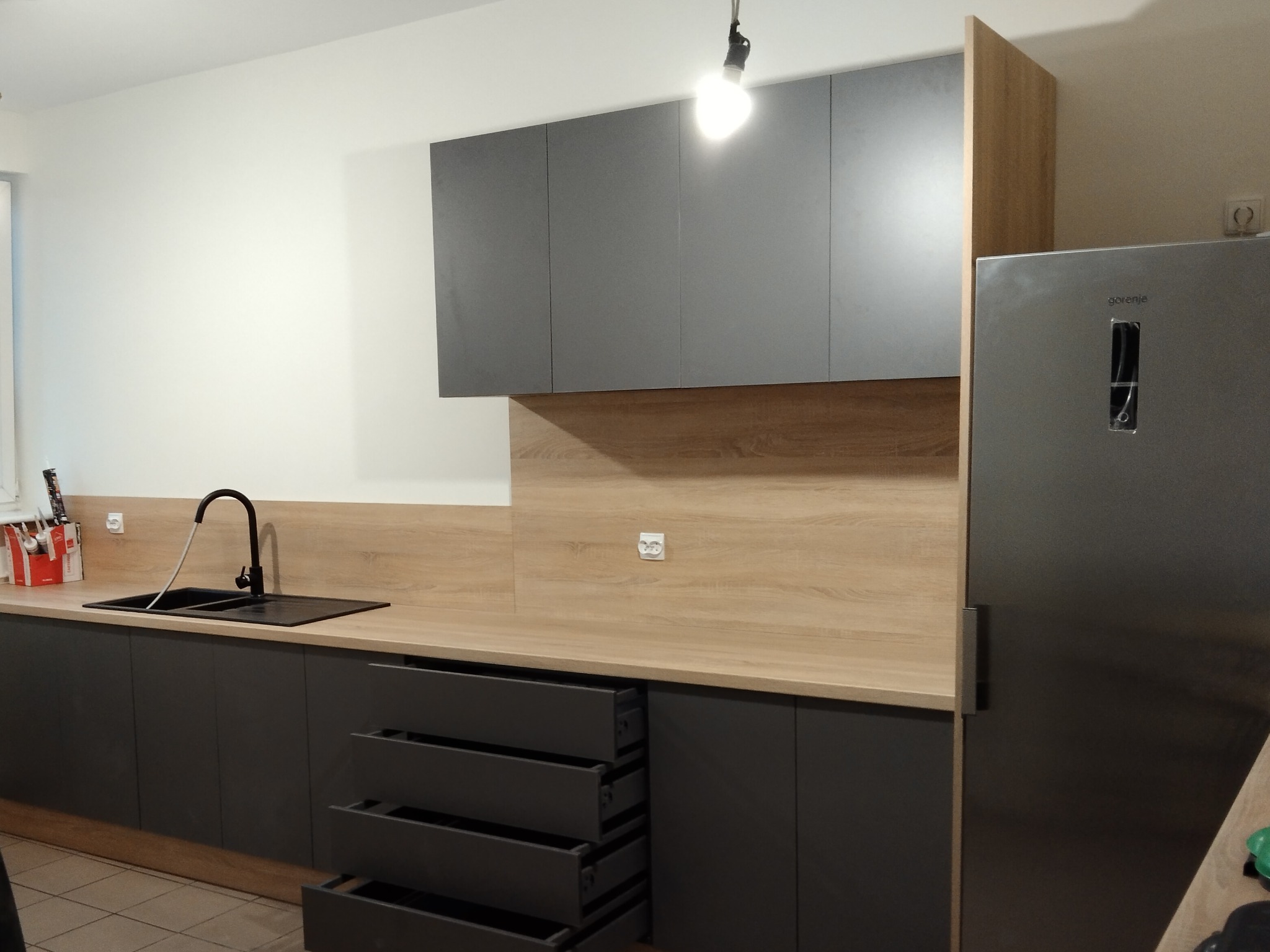 Szara zabudowa kuchenna z drewnopodobnym blatem i otwartymi szufladami. Widoczny zlew z czarną baterią, górne szafki i lodówka. Modernistyczny design.