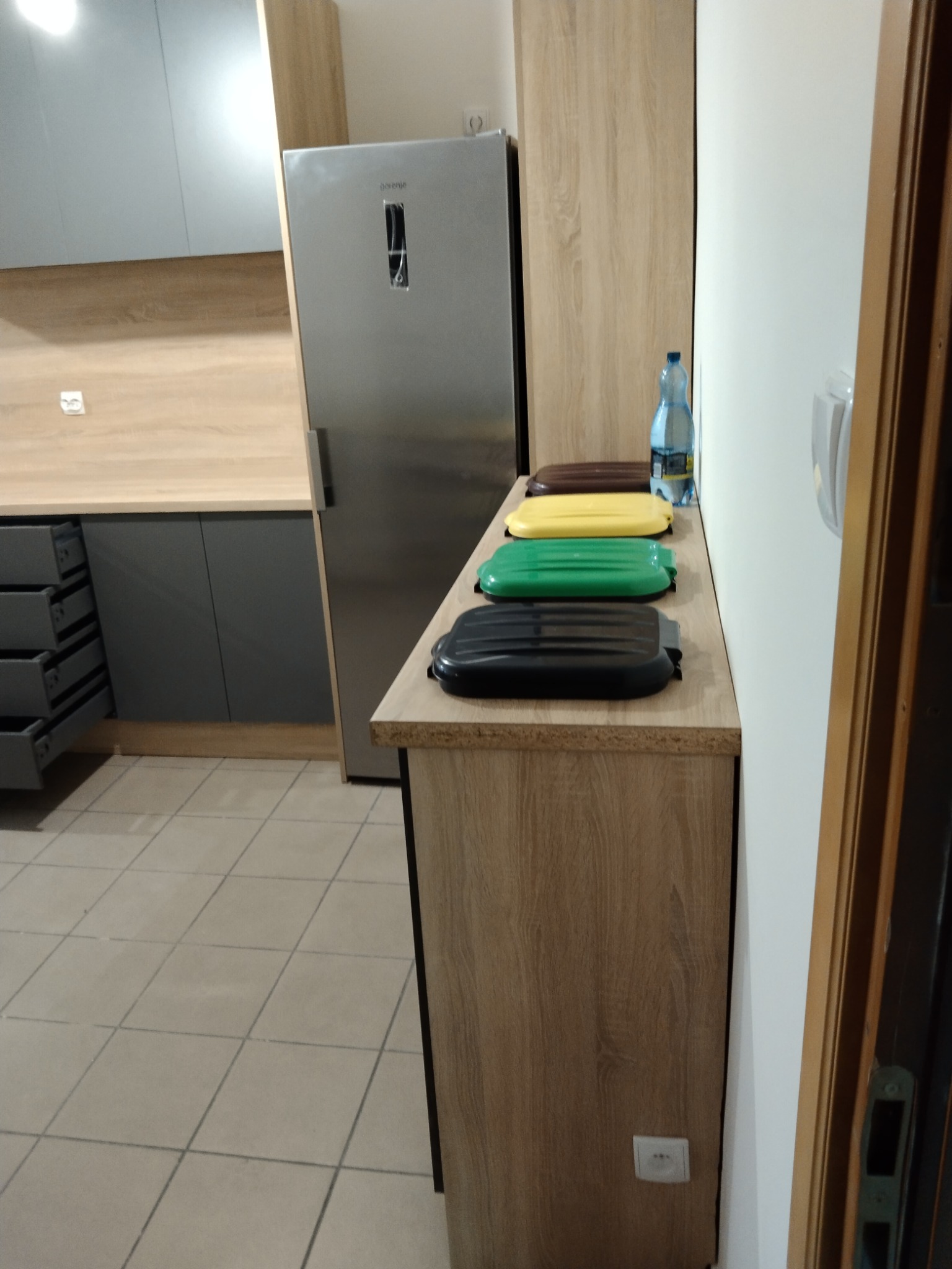 Szare szafki kuchenne z drewnianym blatem, srebrna lodówka i sortownikiem śmieci z kolorowymi pokrywami. Widok na kafelkowaną podłogę i fragment drzwi.