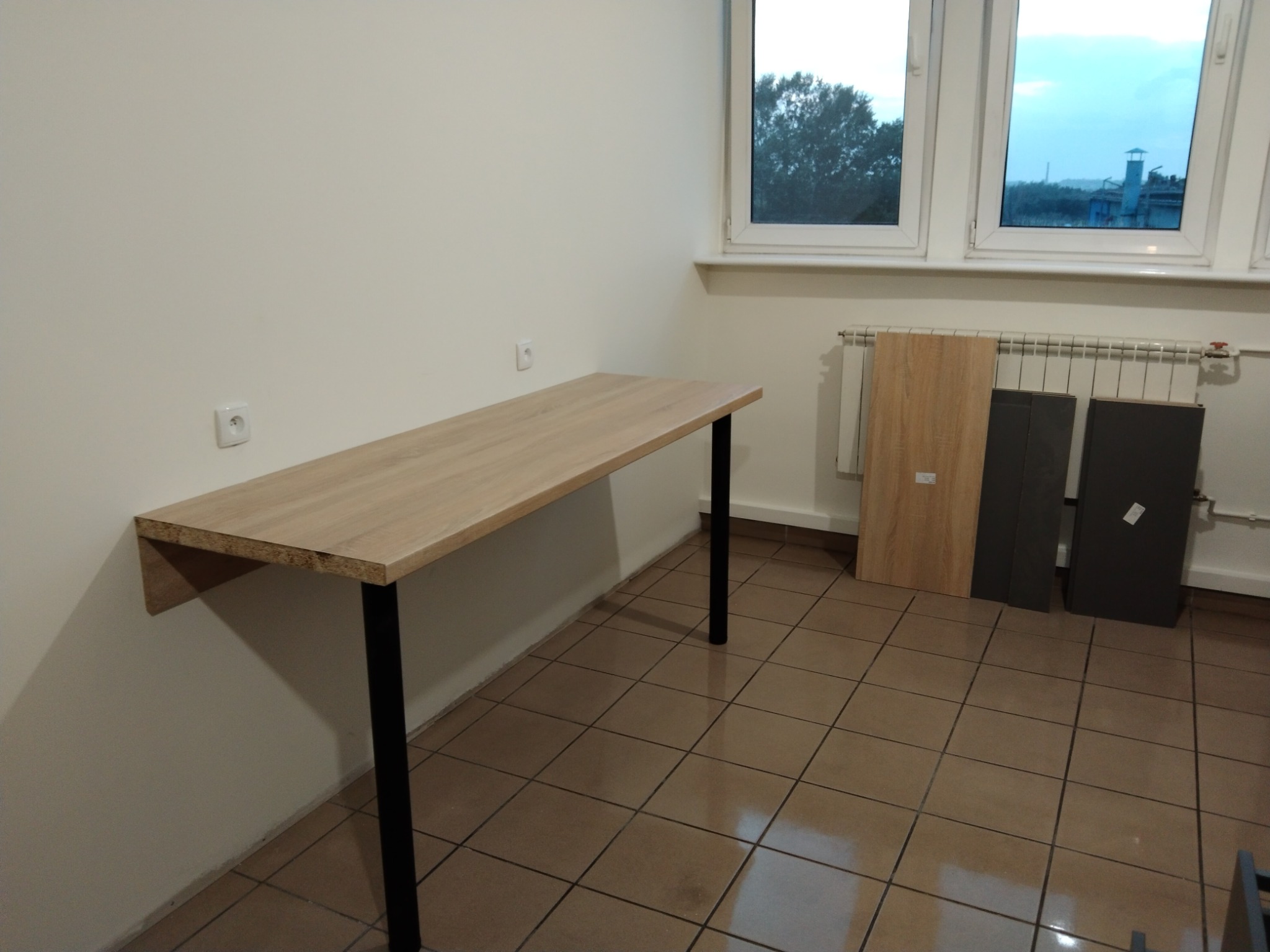 Blat biurka z dębowego drewna, wsparty na czarnych nogach, zamontowany do ściany. W tle okno i grzejnik z panelami. Widoczne gniazdka elektryczne na ścianie.