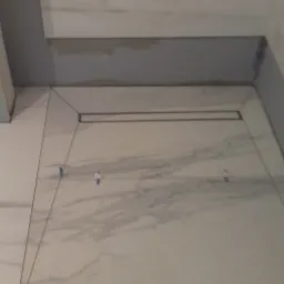 Ułożone białe płytki imitujące marmur z widocznym odpływem liniowym w kabinie prysznicowej, zabezpieczone niebieską taśmą malarską, z szarą ścianą w tle.