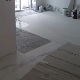 Przygotowanie podłoża pod panele: fragment podłogi z częściowo ułożonymi panelami, zabezpieczona folią strefa i widoczne materiały budowlane w tle, w pokoju z dużym oknem balkonowym.