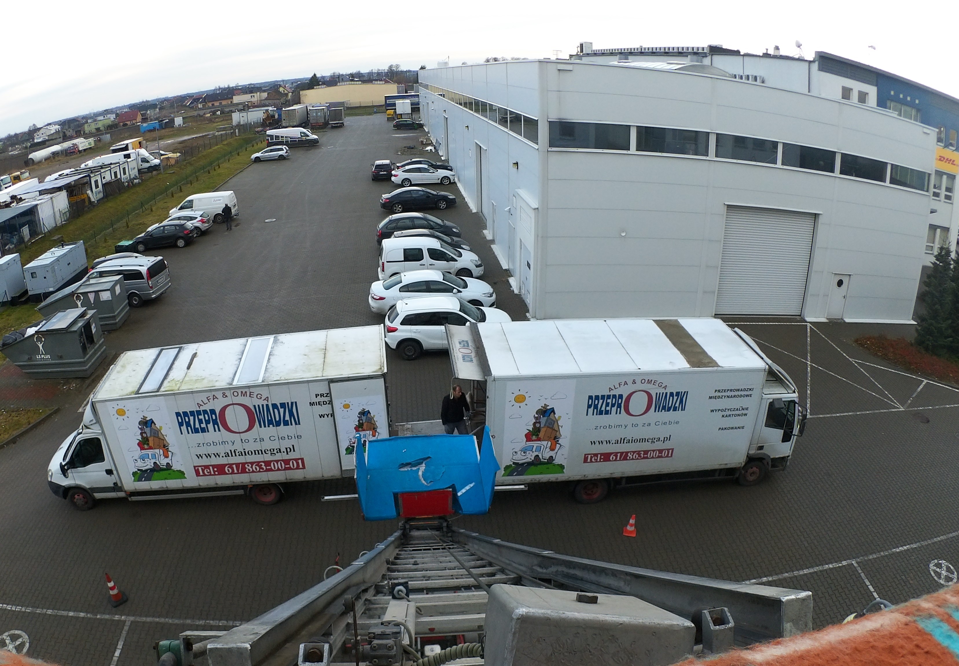 Dwa busy firmy przeprowadzkowej Alfa&Omega z podnośnikiem, na parkingu przed magazynem. Widok z góry z podnośnika. Na busach grafika z transportem rzeczy.