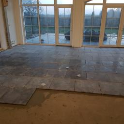 Arkadiusz Jurkowski - Układanie ciemnych, kwadratowych płytek podłogowych, widoczne plastikowe krzyżyki dystansowe i fragment przygotowanego podłoża, duże okna tarasowe w tle.