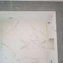 Arkadiusz Jurkowski - Wnęka prysznicowa w trakcie wykańczania, wyłożona białymi płytkami z szarym marmurkowym wzorem, obramowana szarą płytką, widoczne wystające przewody elektryczne.