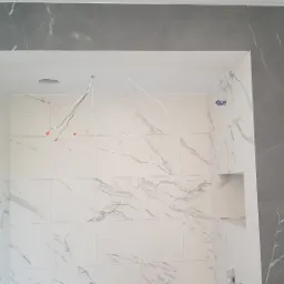 Wnęka prysznicowa w trakcie wykańczania, wyłożona białymi płytkami z szarym marmurkowym wzorem, obramowana szarą płytką, widoczne wystające przewody elektryczne.