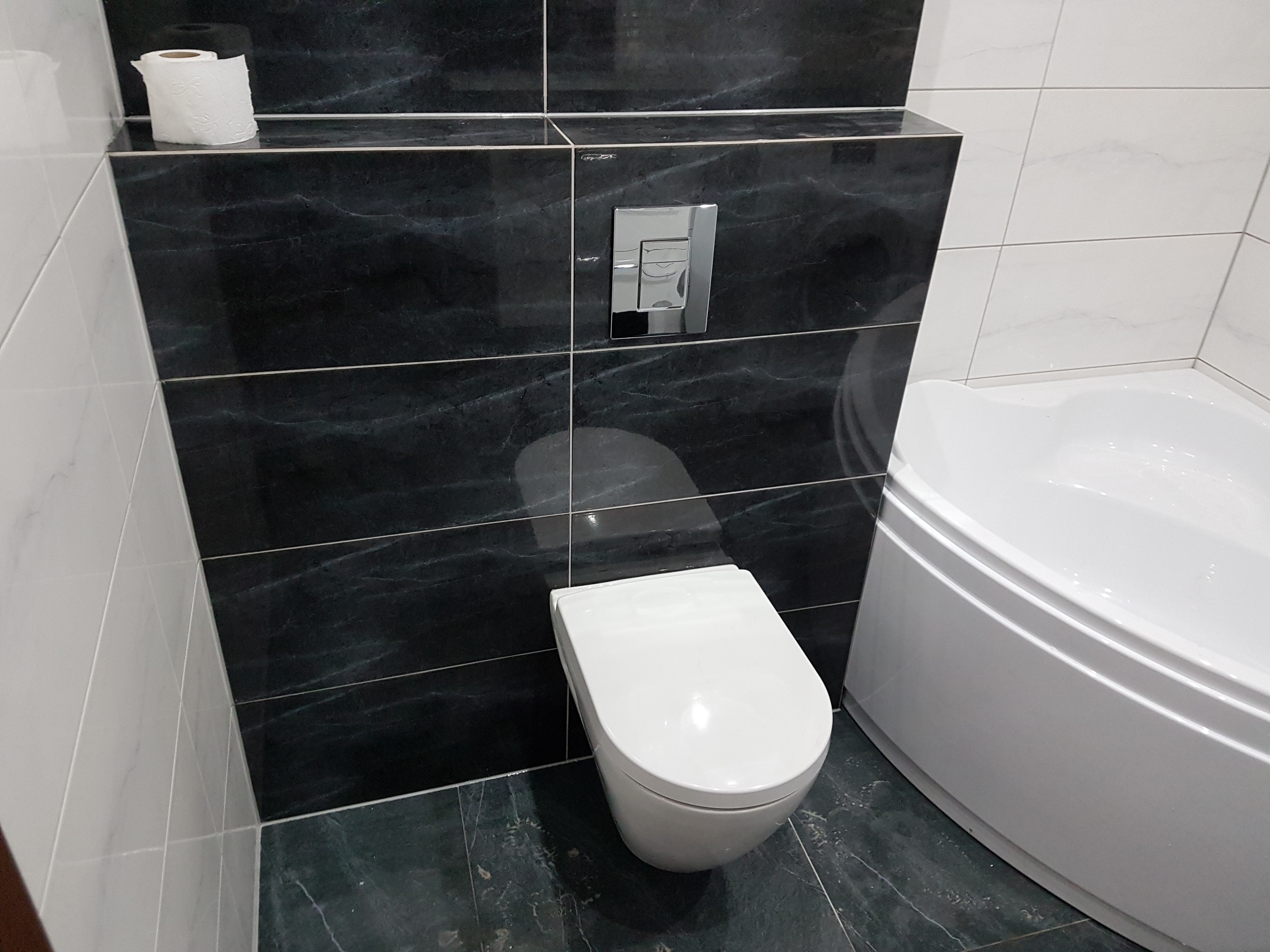 Nowoczesna łazienka z czarno-białymi płytkami marmurkowymi, podwieszaną miską WC z chromowanym przyciskiem spłukującym i narożną wanną.