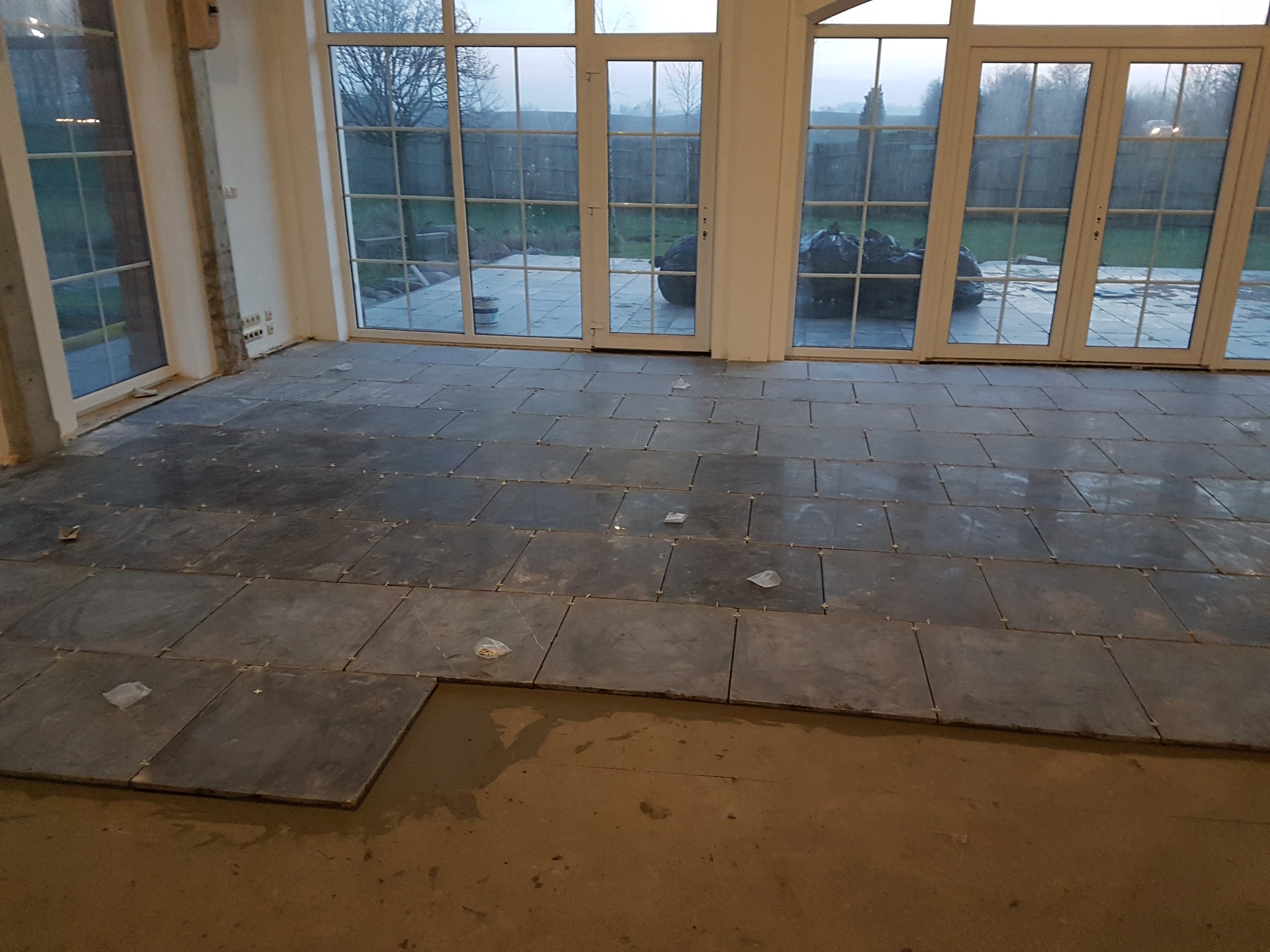 Układanie ciemnych, kwadratowych płytek podłogowych, widoczne plastikowe krzyżyki dystansowe i fragment przygotowanego podłoża, duże okna tarasowe w tle.