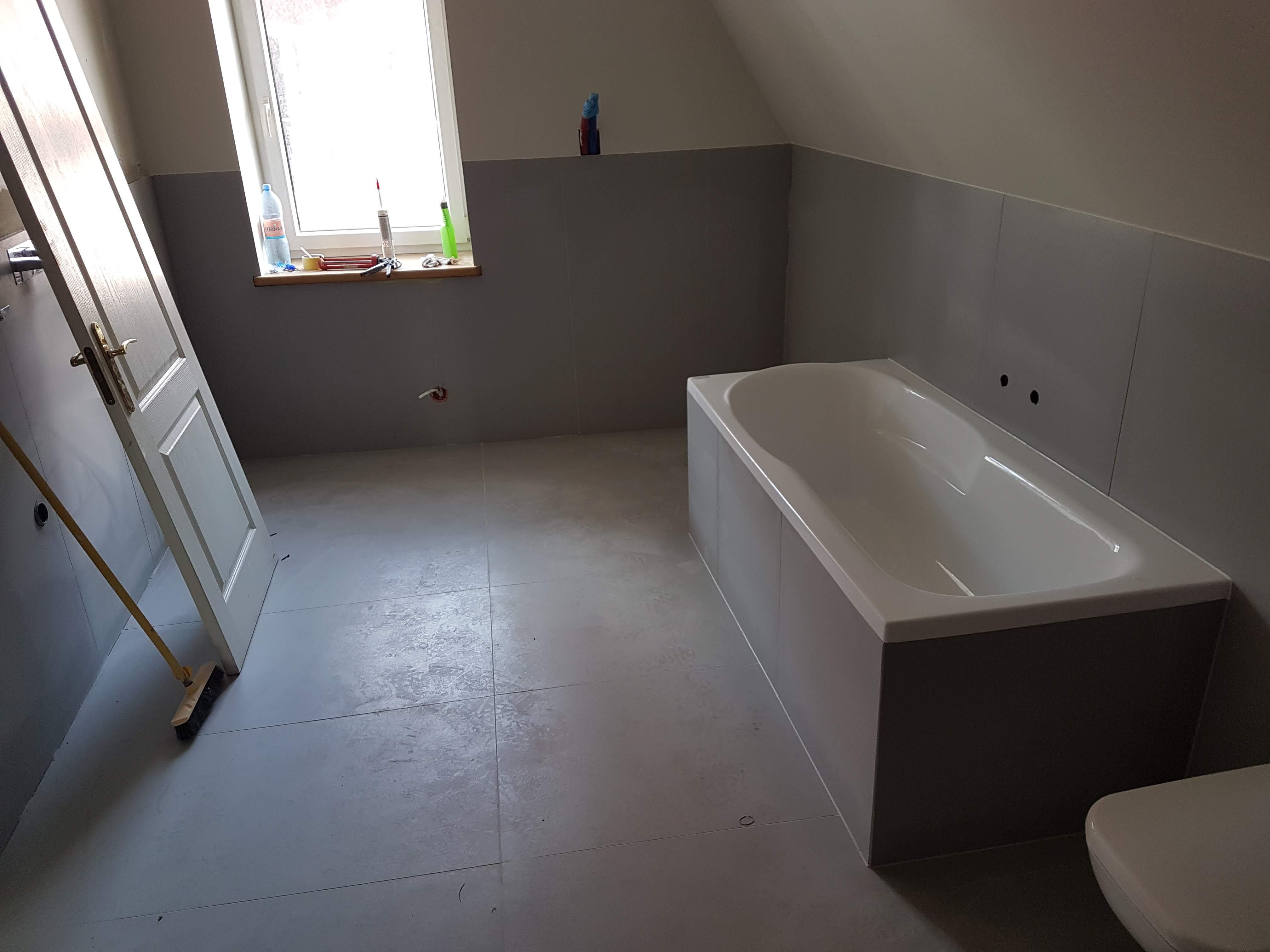 Wnętrze łazienki w trakcie prac wykończeniowych, widoczna wanna obudowana szarymi płytami, szare płytki na podłodze, uchylone białe drzwi z pozłacaną klamką i szczotka oparta o ścianę.