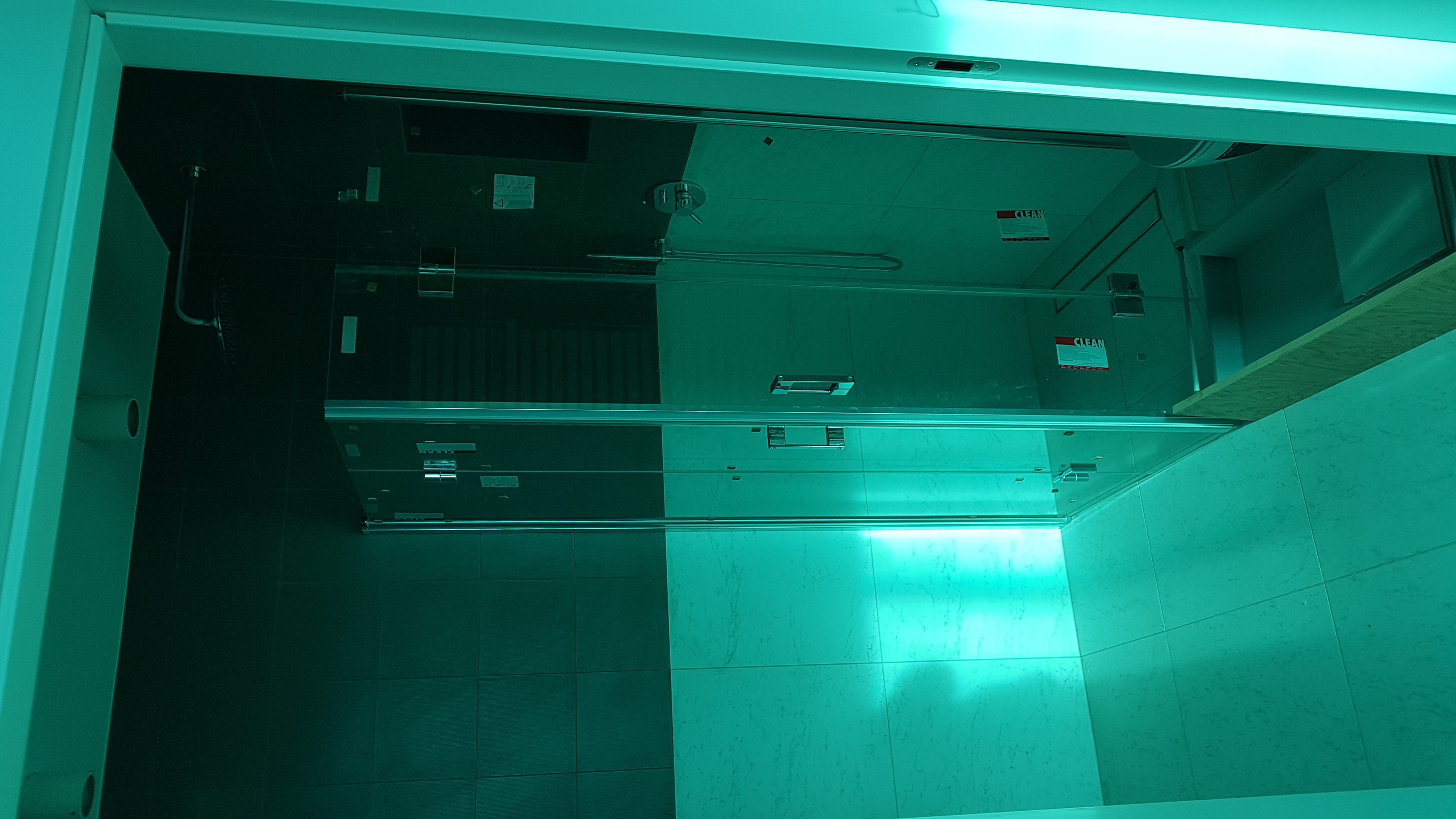 Szklana kabina prysznicowa z chromowanymi elementami, zamontowana w łazience z ciemnymi płytkami, oświetlona intensywnym, jednolitym światłem.