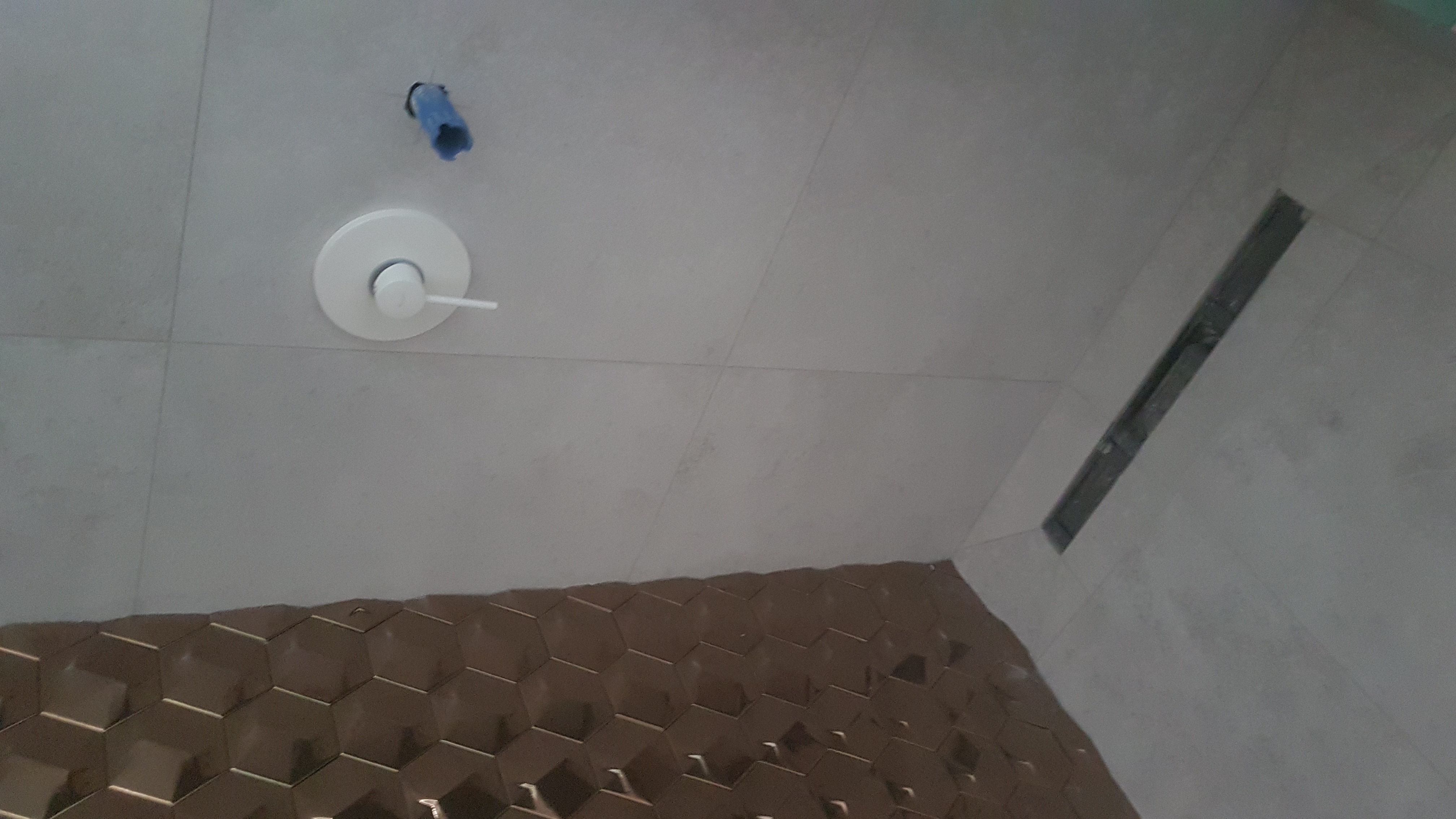 Świeżo wykafelkowana ściana i podłoga w łazience z odpływem liniowym, widoczny zawór baterii podtynkowej i wystający fragment instalacji wodnej.