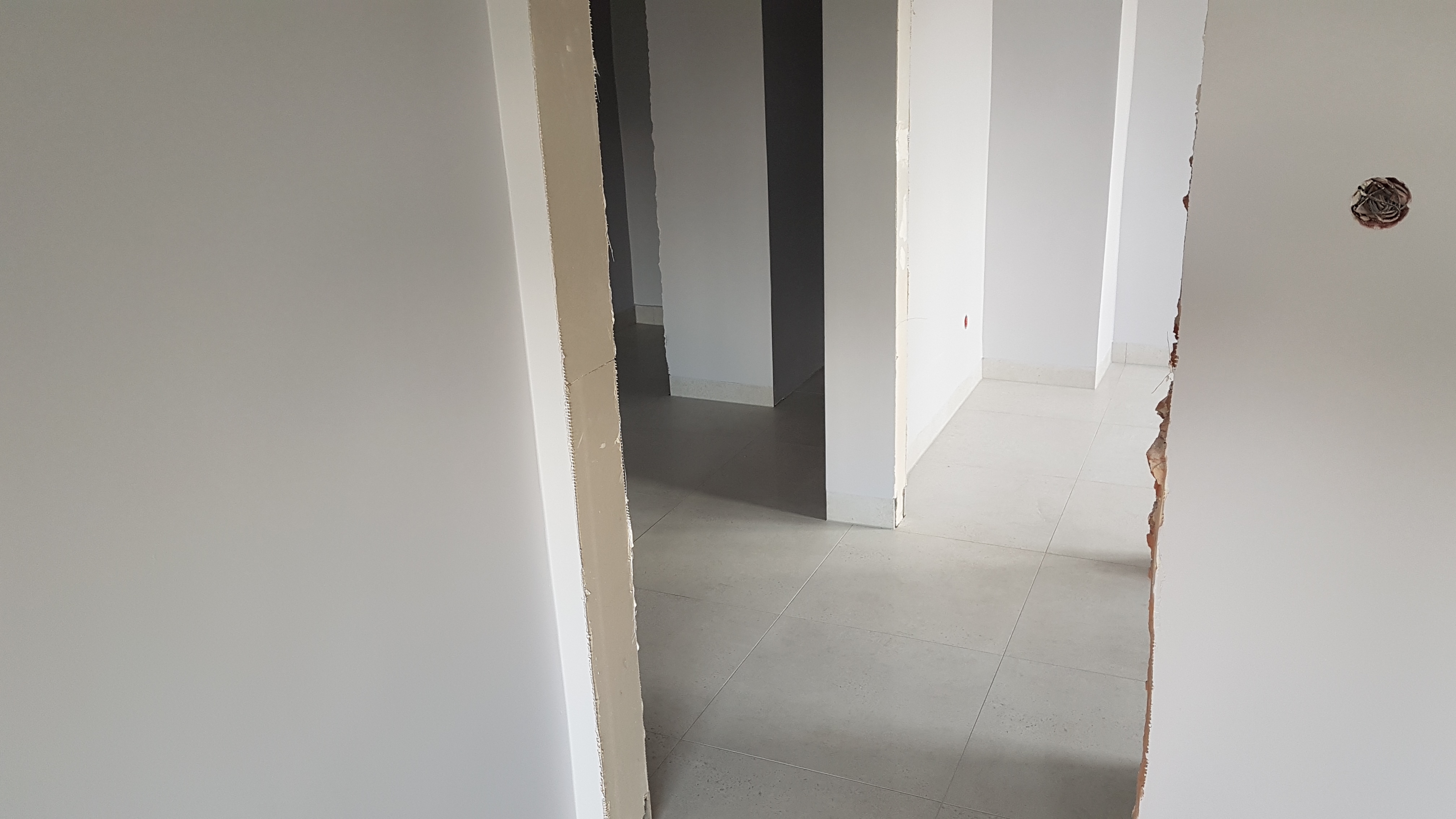 Wnętrze w trakcie remontu: widok przez framugi drzwi na szare płytki podłogowe i białe ściany z widoczną puszką elektryczną i surowymi krawędziami otworów drzwiowych.