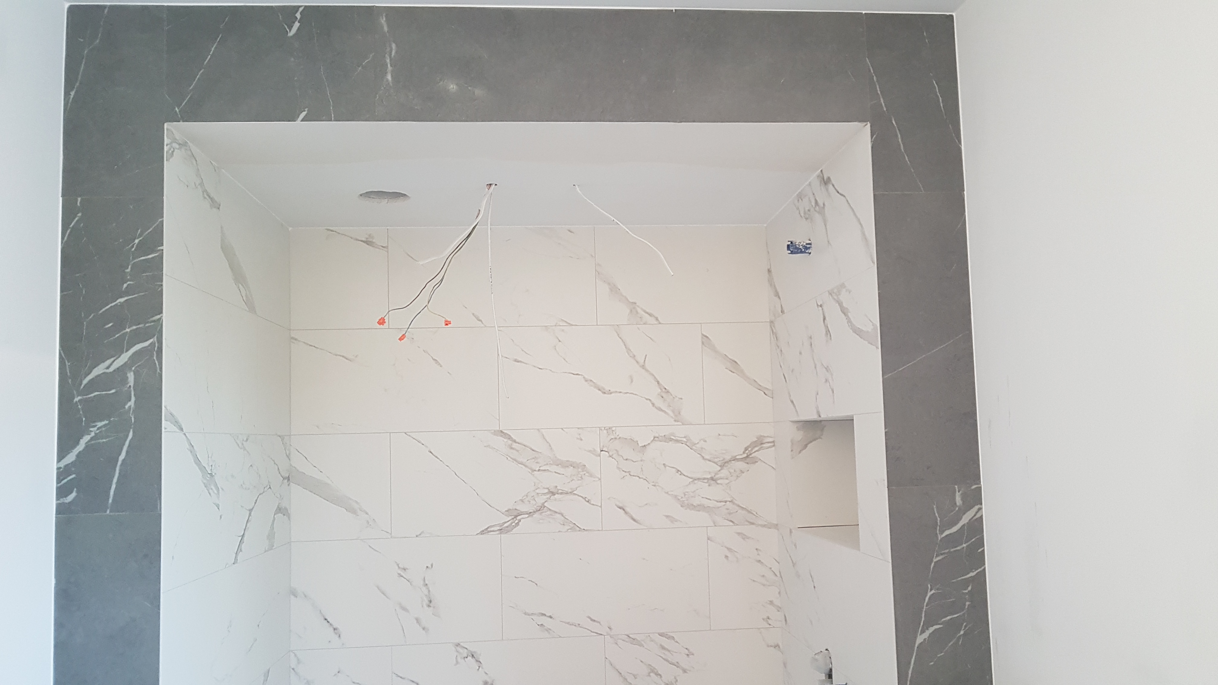 Wnęka prysznicowa w trakcie wykańczania, wyłożona białymi płytkami z szarym marmurkowym wzorem, obramowana szarą płytką, widoczne wystające przewody elektryczne.