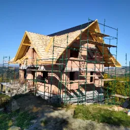 Dom w trakcie budowy z częściowo położonym pokryciem dachowym, widoczne rusztowania i drewniana konstrukcja dachu, krajobraz w tle.