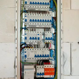 Rozdzielnica elektryczna Schrack zamontowana na ścianie z białego bloczka, widoczne zabezpieczenia różnicowoprądowe, bezpieczniki, przewody w kolorach żółto-zielonym, niebieskim i brązowym,...
