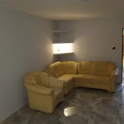 Jasny narożnik i fotel w salonie z szarą podłogą imitującą beton, białe ściany z p&oacute;łkami podświetlonymi od dołu, osoba sprzątająca w tle.