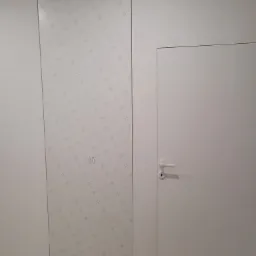 Białe, minimalistyczne drzwi ukryte w ścianie, obok panel ścienny z delikatnym wzorem, tworzące spójną, nowoczesną kompozycję.