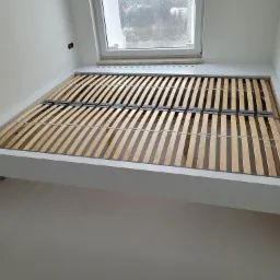 Rama łóżka z drewnianym stelażem, wbudowana w białą wnękę pod oknem, widok z dołu.