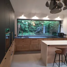 Nowoczesna kuchnia z drewnianymi frontami szafek i dużym oknem z widokiem na zieleń ogrodu, oświetlona designerską lampą sufitową i punktowymi halogenami.